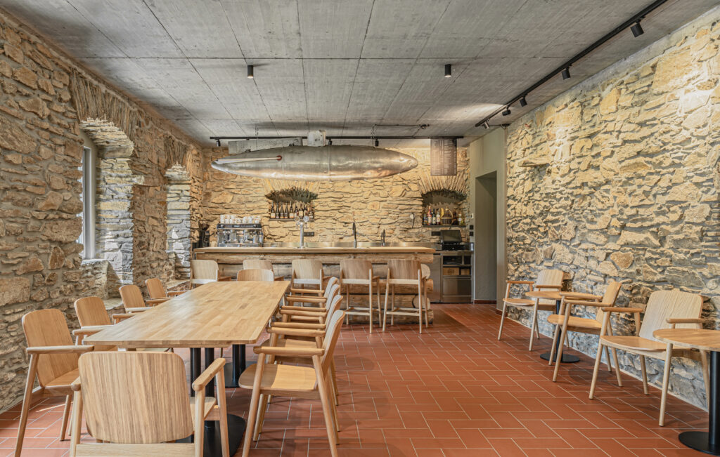 Dřevěné stoly a židle v interiéru restaurace U Pašeráka v Suché. V pozadí bar z masivních kmenů. Návrh: ateliér Orchitekti.